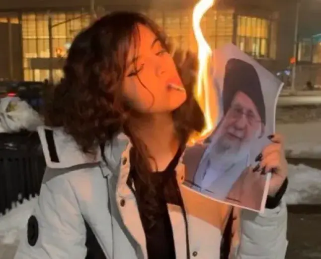 a woman lighting a cigarette with a burning phot of the Ayatollah