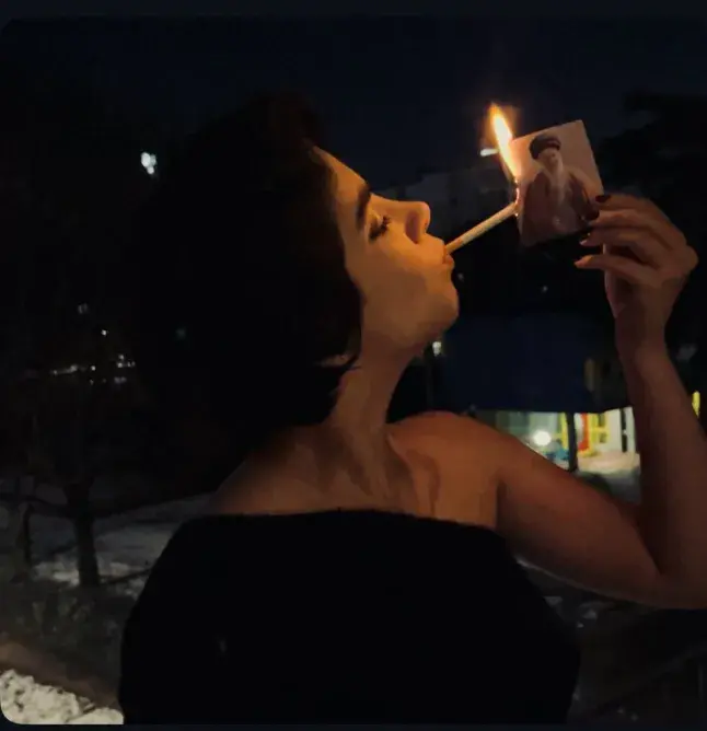 a woman lighting a cigarette with a burning phot of the Ayatollah