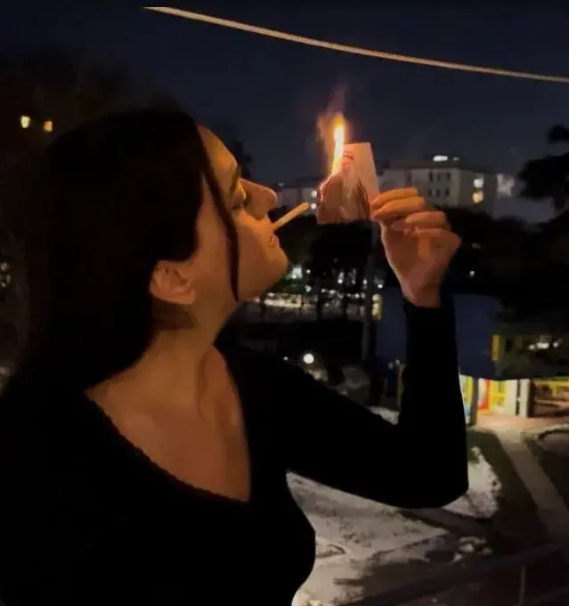 a woman lighting a cigarette with a burning phot of the Ayatollah