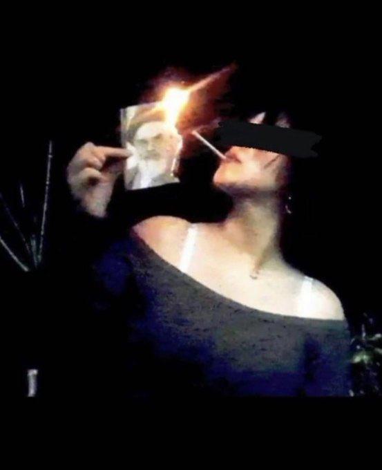 a woman lighting a cigarette with a burning phot of the Ayatollah