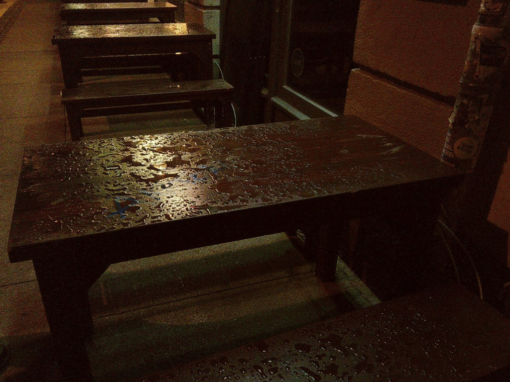 An evening street scene. Dark brown wooden benches and tables next to a wall. Raindrops and larger puddles of water on these.