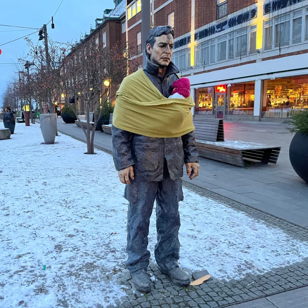 A statue of a man standing in a snowy urban plaza is wrapped with a yellow baby sling holding a doll wearing a pink hat. This is part of a campaign by Arena Idé for International Men’s Day, aimed at addressing unequal parental roles in Sweden. The use of the baby sling symbolizes fatherhood and caregiving, highlighting the need for greater participation of fathers in childcare and challenging societal norms about gender roles. The surrounding area features benches, shops, and evening lighting, emphasizing the public and thought-provoking nature of the campaign.