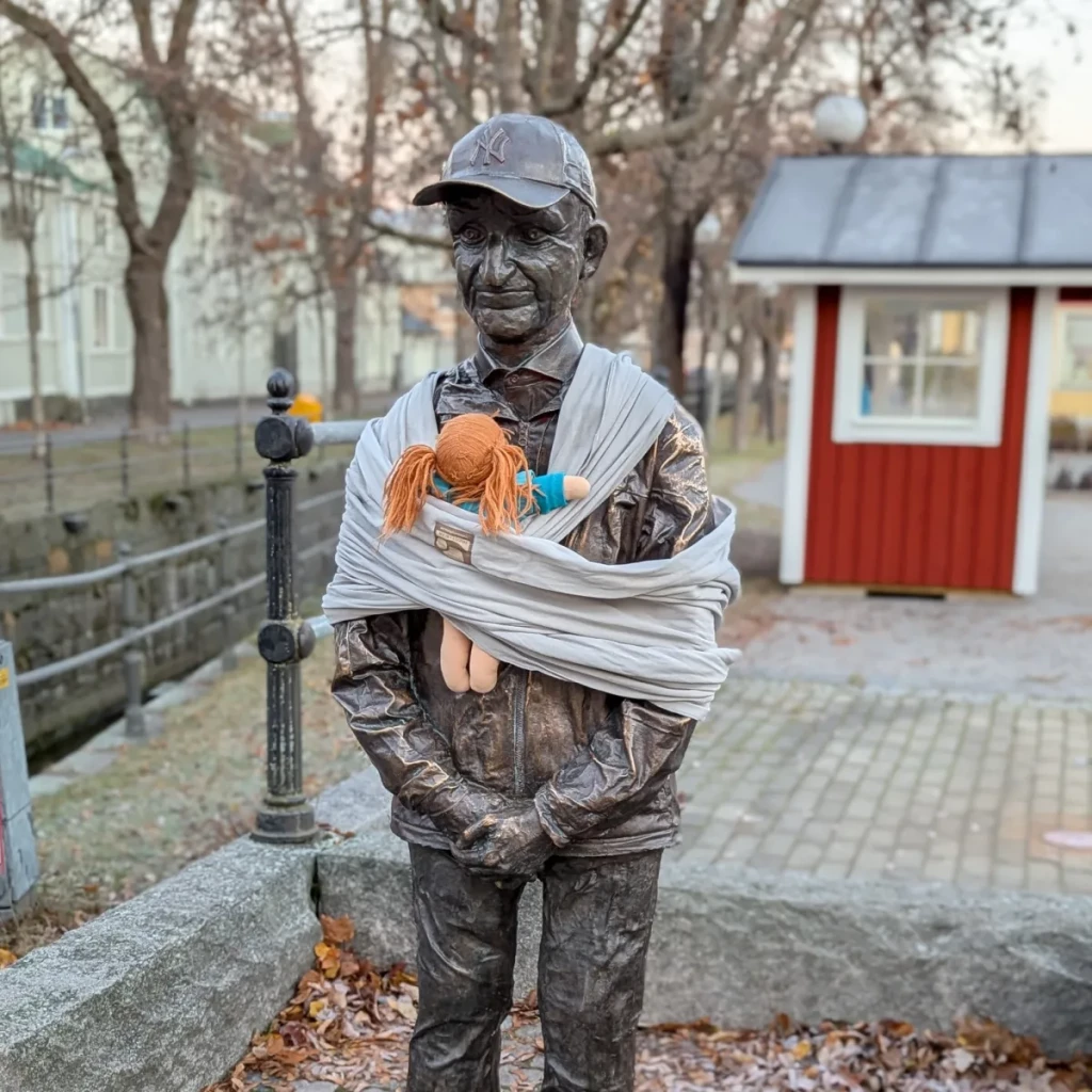 A life-sized bronze statue of a man wearing a baseball cap is adorned with a light gray baby sling, holding a doll with orange pigtails and colorful clothes. This installation is part of Arena Idé's International Men’s Day campaign, which aims to raise awareness about the unequal distribution of parenting responsibilities in Sweden. The statue is placed in an outdoor setting with autumn leaves, a red wooden building, and railings in the background, blending everyday life with the campaign's thought-provoking message on fatherhood and shared caregiving roles.