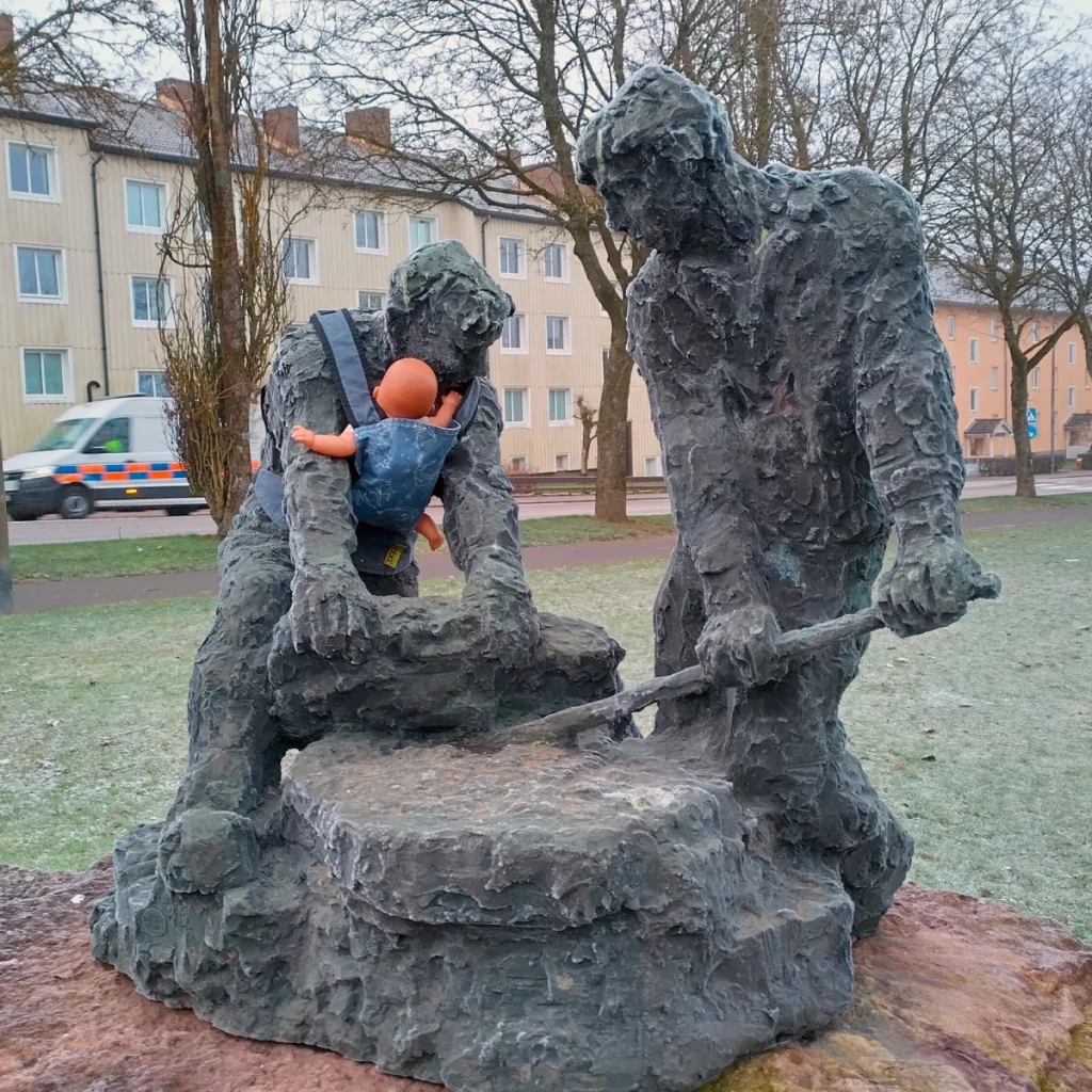 A bronze statue of two men working together on a large grinding stone is modified with a baby carrier strapped to one of the figures, holding a doll. The baby carrier, a modern addition, contrasts with the rugged, labor-intensive depiction of the figures. This intervention is part of Arena Idé's International Men’s Day campaign, drawing attention to the unequal sharing of parenting responsibilities in Sweden and encouraging societal reflection on the role of fathers. The backdrop includes residential buildings, a frosty ground, and a passing vehicle, situating the scene in a contemporary urban context.