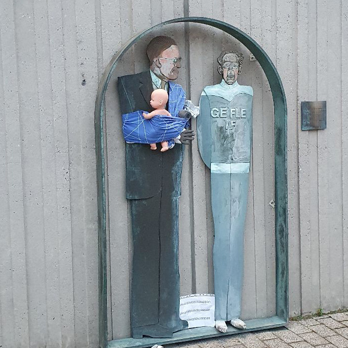 A painted metal statue of two figures, one in a suit and the other in a sports uniform labeled "Gefle IF," is modified with a blue baby sling holding a doll. The sling, wrapped around the figure in the suit, represents a modern caregiving role. This creative adjustment is part of Arena Idé's International Men’s Day campaign, addressing the unequal sharing of parenting duties in Sweden. Set against a concrete wall with an arched frame and informational signs below, the scene highlights the campaign's aim to challenge traditional gender roles and advocate for shared parental responsibilities.