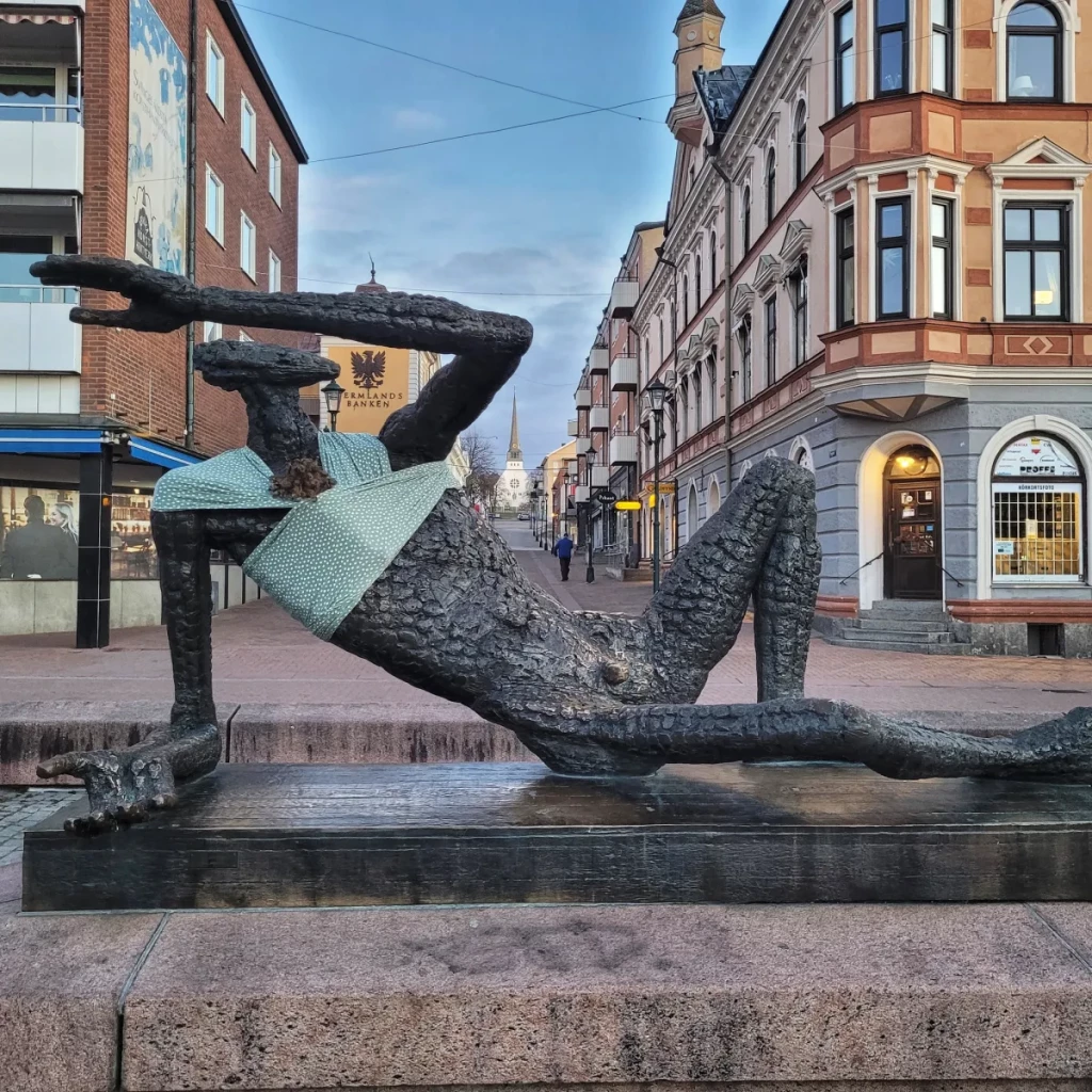 A modern bronze statue of a reclining figure, with textured skin and an abstract design, is humorously adorned with a light green baby wrap. The wrap, draped around the statue's upper body, transforms the figure into a symbolic participant in caregiving, aligning with Arena Idé's International Men’s Day campaign. Set in a busy urban street surrounded by historic buildings, the installation challenges societal norms and promotes discussion about the unequal distribution of parenting responsibilities in Sweden, emphasizing the importance of shared caregiving roles.