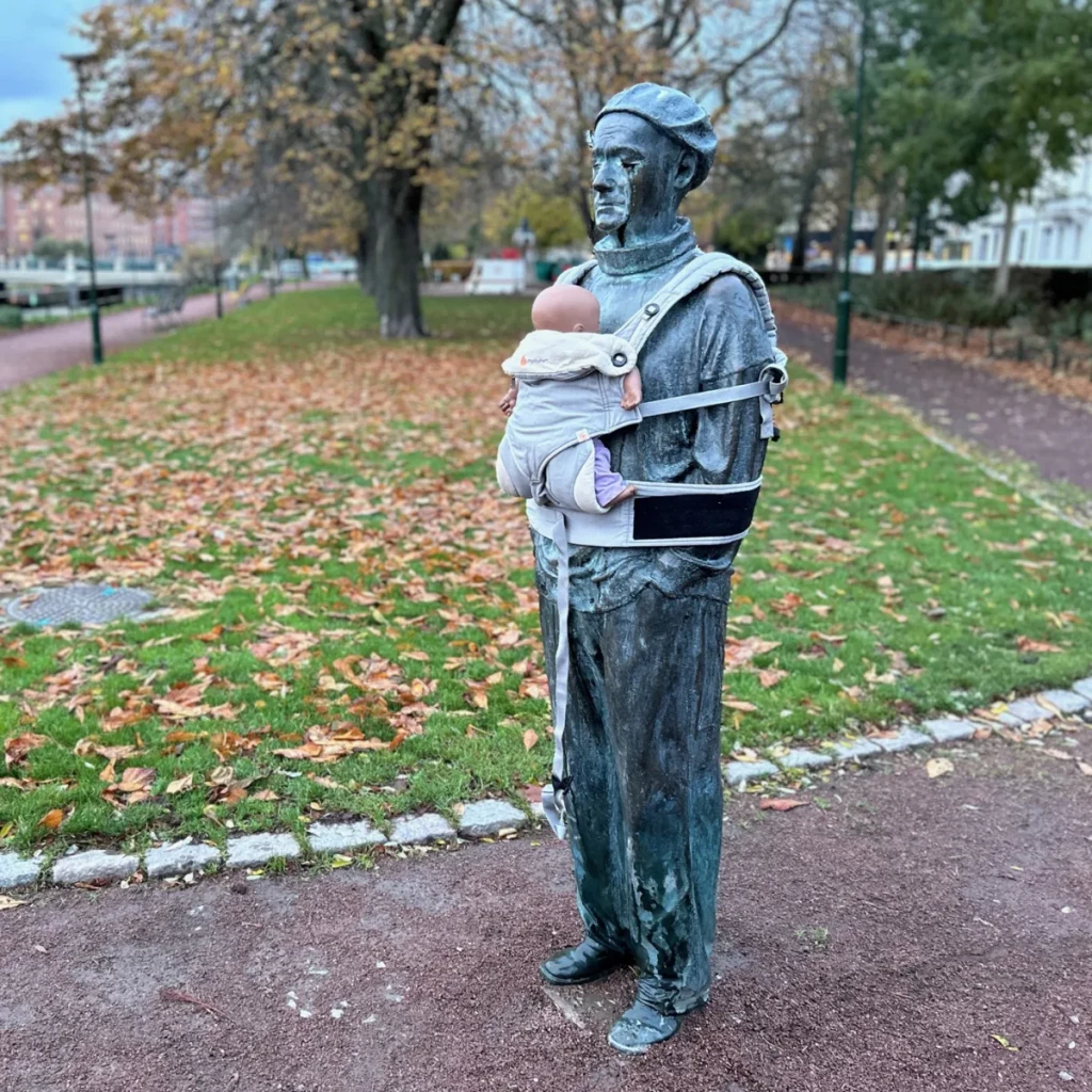 A bronze statue of a man wearing a beret is fitted with a modern baby carrier holding a doll. The carrier, secured to the figure's chest, contrasts with the statue’s traditional stoic demeanor, symbolizing the caregiving role of fathers. This artistic intervention is part of Arena Idé's International Men’s Day campaign, aiming to spotlight the unequal distribution of parenting responsibilities in Sweden. Set in a park with fallen autumn leaves, trees, and walking paths, the installation invites reflection on the importance of shared parenting in both work and family life.