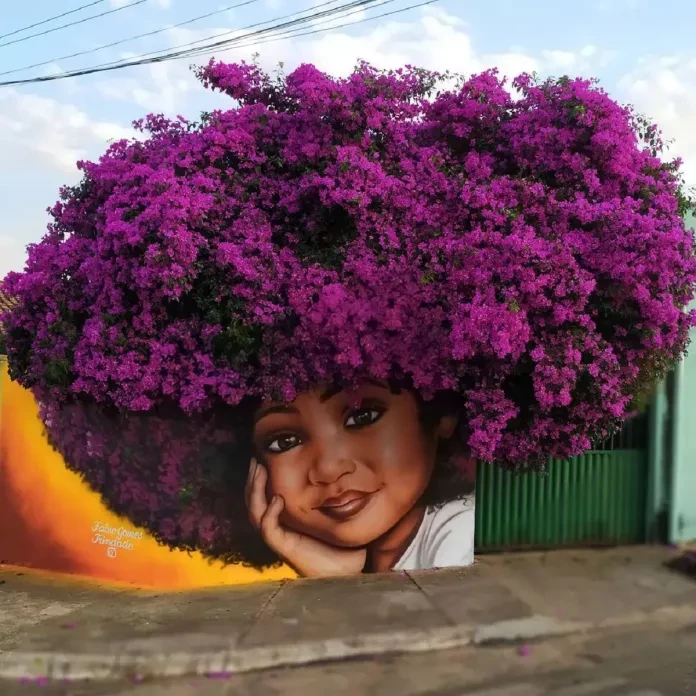 A stunning mural by Fábio Gomes Trindade in Goiás, Brazil, depicting the face of a young Black girl with a radiant smile, her resting hand supporting her chin. The girl's afro is seamlessly integrated with vibrant, blooming magenta bougainvillea flowers from a nearby tree, creating a breathtaking blend of natural and painted elements. The backdrop features warm orange and yellow tones, enhancing the contrast and warmth of the artwork.