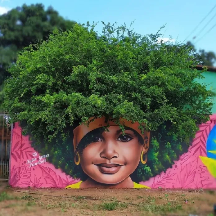 A beautiful mural by Fábio Gomes Trindade in Goiás, Brazil, portraying a young Black girl with a confident smile, framed by a vibrant green tree that forms her afro. The girl wears a yellow headband and hoop earrings, with pink floral patterns on the wall background. The fusion of natural greenery and painted art creates a harmonious and striking visual.