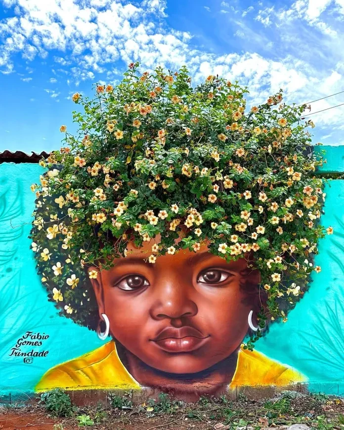 A vibrant mural by Fábio Gomes Trindade in Goiás, Brazil, showcasing a young Black girl with a calm, steady gaze. Her afro is creatively represented by a lush bush of yellow and orange blossoms, blending nature seamlessly with the painted portrait. The turquoise wall background and clear blue sky add a refreshing contrast, making the artwork radiate warmth and vitality.