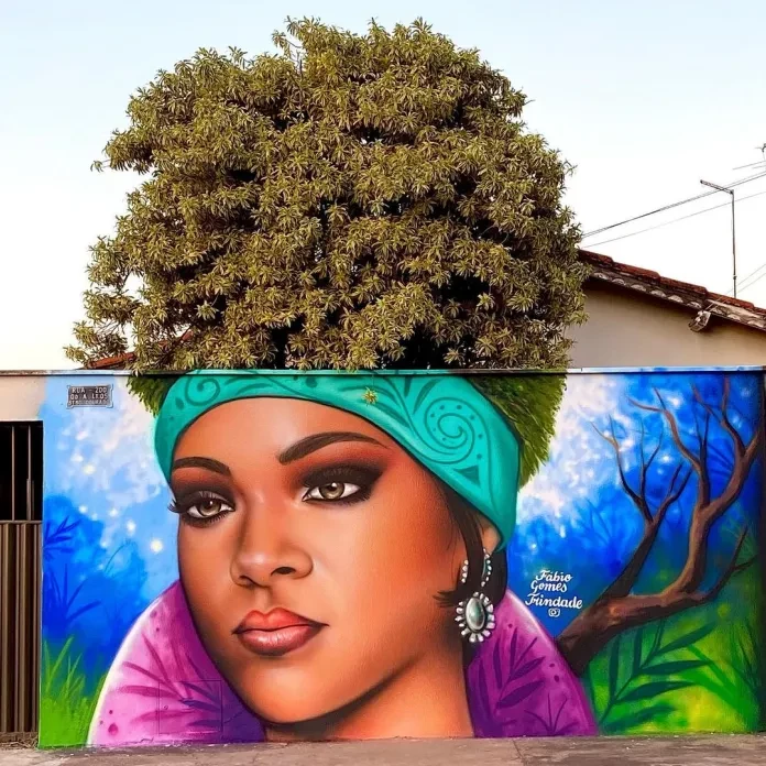 A stunning mural by Fábio Gomes Trindade in Trindade, Brazil, honoring Rihanna. The artwork features a detailed portrait of Rihanna with her afro seamlessly integrated into the lush greenery of a large tree above. The vibrant background includes blue skies and nature-inspired elements, perfectly blending art and the environment.