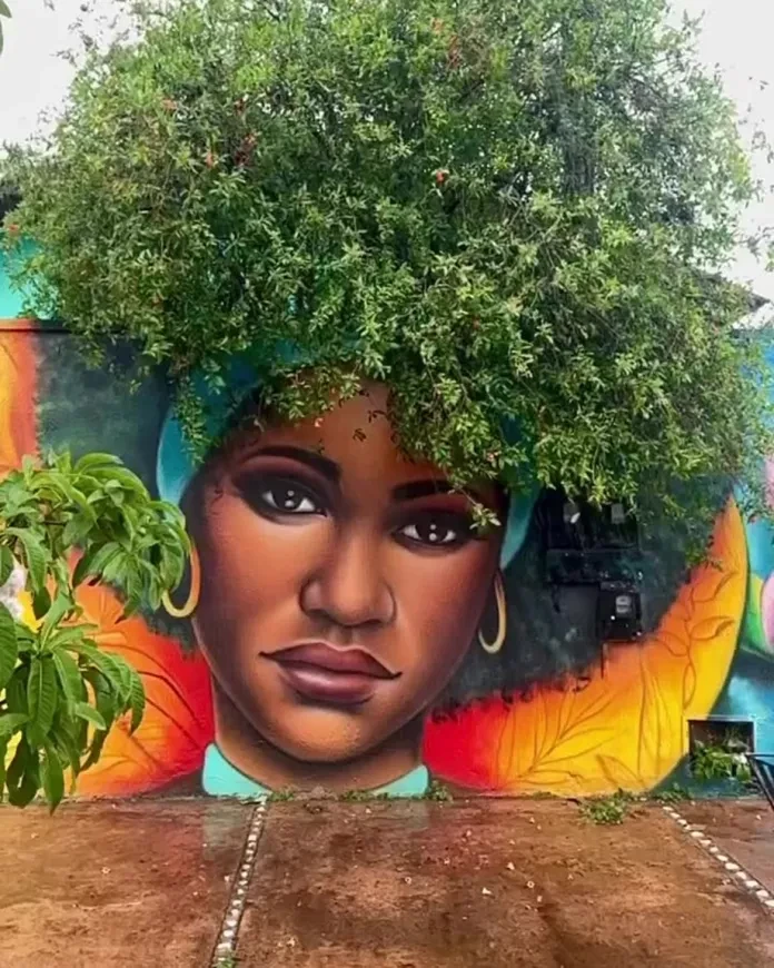 A mural by Fábio Gomes Trindade in Trindade, Brazil, featuring the portrait of a Black woman with a serene expression. Her afro is creatively extended using the lush foliage of a tree above the wall. The vibrant background includes orange and teal hues, blending the artwork harmoniously with its environment.