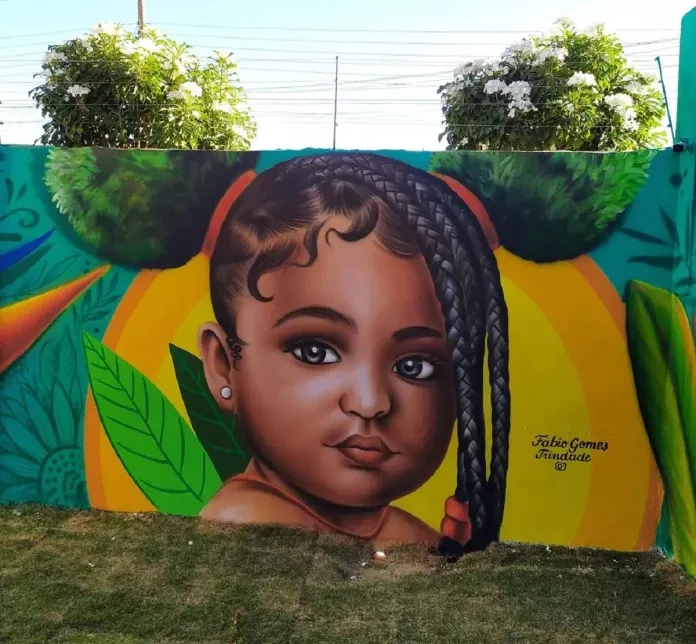 A vibrant mural by Fábio Gomes Trindade in Trindade, Brazil, featuring a portrait of a young child with braided hair and gentle curls framing their face. The background incorporates vivid tropical elements, such as green leaves and orange shapes, with the afro creatively extended using lush greenery above the wall. The bright colors and intricate details create a lively, harmonious blend of nature and art.