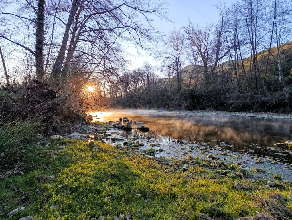 Photo d'un bord de petit fleuve. À l'avant-plan, on voit une berge couverte de gazon vert et parsemée de petites pierres. La rivière reflète la lumière du soleil, qui se lève entre les arbres dénudés. Une légère brume s'élève au-dessus de l'eau calme. À l'arrière-plan, on devine un petit massif karstique.