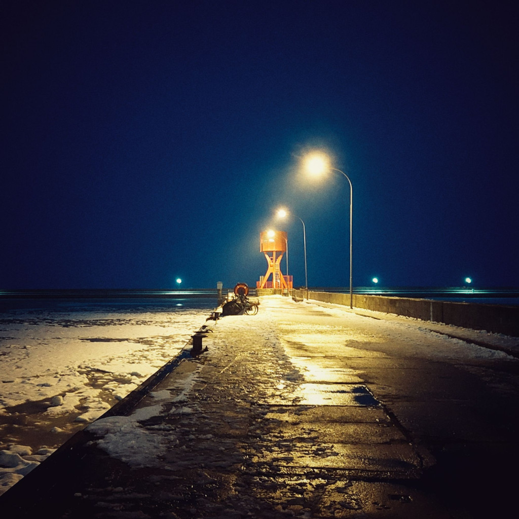 A quay in night light. Icy water on the left. A red lighthouse in the middle.