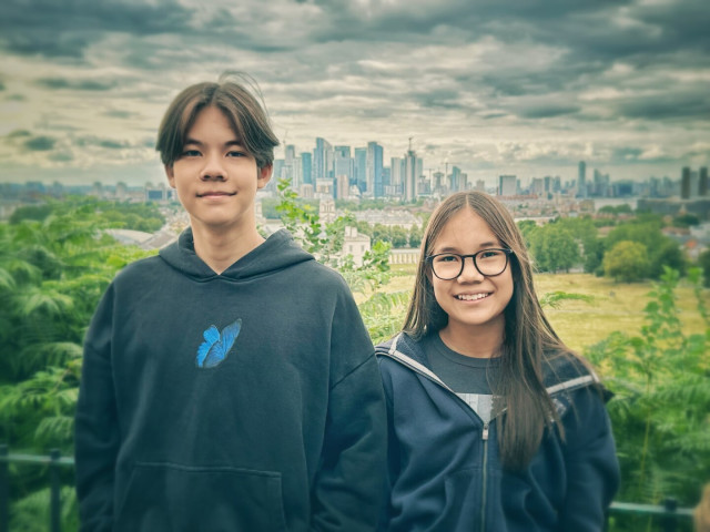 Two teenagers posing for photo. They are on the hill of Greenwich, London, with a park and a cityscape in the background.