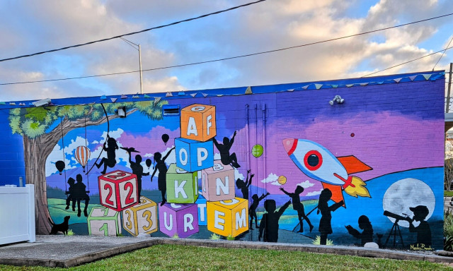The side of a modest pre-school building colorfully painted with a mural showing the silhouettes of children exploring science, art, and learning. With building blocks,  space ships, and nature scenes.