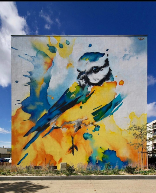 Streetartwall. A mural of a beautiful colorful bird was sprayed/painted on the exterior wall of a four-story modern building. A yellow and blue bird sits in the middle of an explosion of bright colors. A mural that looks like a giant watercolor and adds a great splash of color to this uniform large housing development.
(In front of the mural is a beige sidewalk and above it is a blue cloudy sky to match the mural.)