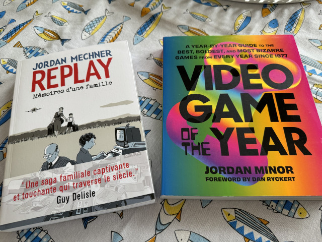 Two books on a table with a fish-patterned tablecloth. The left book is titled "REPLAY" by Jordan Mechner and the right is "VIDEO GAME OF THE YEAR" by Jordan Minor.