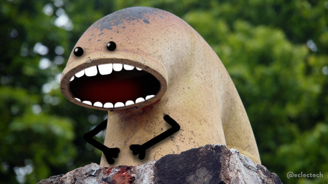 The photo is of a pale orange-yellow pipe on top of a stone pillar backed by a soft focus green leaf canopy of some large trees. The pipe is curved towards the foreground with an oval opening, and behind splits into two down-pipes that disappear into the stone. Two eyes have been drawn above the opening, and teeth have been added to turn it into a mouth. Two arms rest on what would be its belly if it has one.