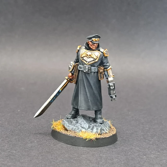 An Imperial Guard Commissar in a black coat and hat. He is holding a bolt pistol in one hand and a power sword in the other