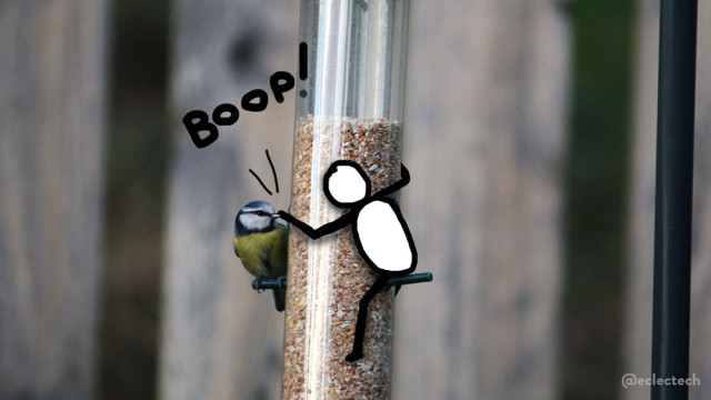 Photo of a bird feeder, partly filled with seed, with soft focus wood fencing in the background. A blue tit sits on one of the feeding perches, looking towards the feeder. On the opposite perch a simple drawn figure sits, reaching round to boop the bird on the beak. The work BOOP! is written above.