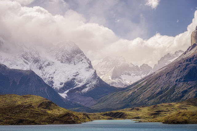 Jagged snow-capped peaks rise dramatically above Lake Pehoe in Torres del Paine National Park in Chile. Lush green hills roll gently in the foreground under a cloudy sky.