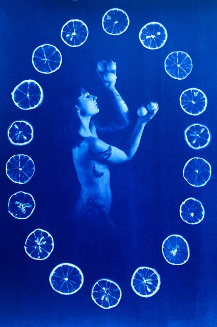A naked woman is standing sideways, holding lemons in her hands. The image is framed in an oval shape made by lemon slices. Blue and white. 