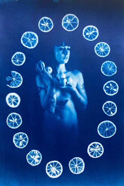 A naked woman is standing to the camera, holding lemons in her hands. The image is framed in an oval shape made by lemon slices. Blue and white. 