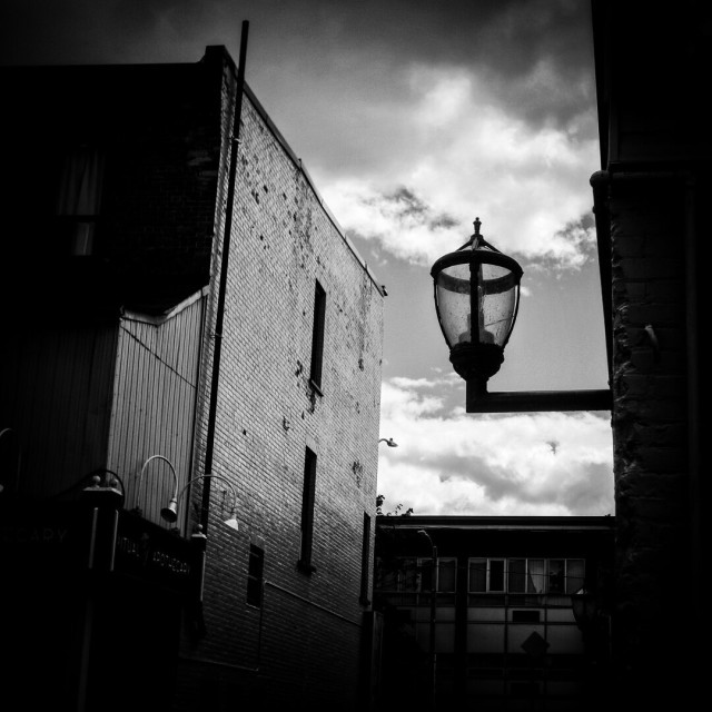 A glass light fixture attached to the wall of a building is seen against the bright sky in this dark black and white image.