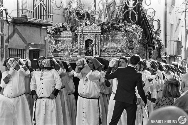 A black-and-white film photo of a large float carried in a Holy Week procession through the streets. There are probably 50 or more people carrying this float. A man in a black suit coordinates the carriers and guides them along the narrow streets.