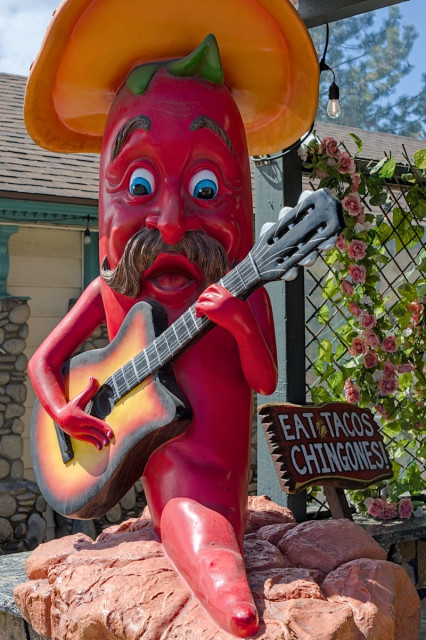 A large human size statue of a red chile pepper with a human face, a dark mustache, and sombrero playing a guitar. He sits on a rock. To his right is a small sign that says, "Eat Tacos Chingones!"