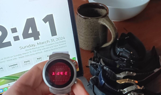 A hand holding an LED watch, showing the seconds, currnetly "41". In the background is a computer screen also showing the time and date. The seconds show "41". To the right of this is a pile of watches and a coffee cup.