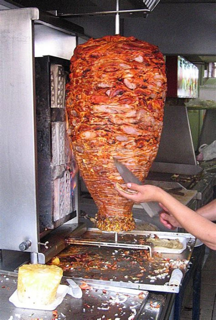 A large conical shaped piece of meat hangs from a spit over a chrome pan. The meat spins on the spit and is cooked by an electric coil. Somebody's arm are visible. One hand holds a knife and the other has a tortilla cup in the palm. The knife is shaving meat off of the spinning roast into the tortilla. A large piece of pineapple sits on the counter in the lower left of the frame.