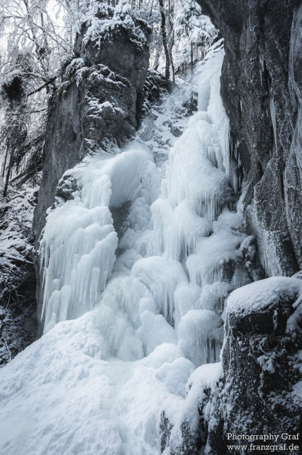 This captivating image showcases the serene beauty of a frozen waterfall nestled amidst a winter landscape. The scene is enveloped in a blanket of snow, emphasizing the season's quiet and peaceful ambiance. Dominated by shades of grey and white, the photograph presents a harmonious blend of the winter's chill and nature's grace. The waterfall, now a sculpture of ice, becomes the central figure, surrounded by snow-covered trees that stand as silent guardians of this secluded spot. The stream that once cascaded freely is now stilled, encapsulated in ice, contributing to the scene's frozen tranquility. The landscape, a testament to the season's power to transform, is a mesmerizing view of winter's cold embrace, where the natural world pauses in a state of suspended animation. This image, a celebration of the season’s stark beauty, invites viewers to marvel at the quiet majesty of a winter's day, where nature's movements slow, and its landscapes are reimagined in ice and snow.