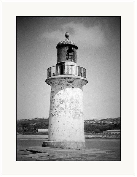 Grainy black and white film photograph showing an old lighthouse at the end of a pier.  It looks weathered and old, the glass at the top is broken.