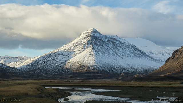 A photo of a mountain in daylight. The sun is on the left of the shot and highlights the flanks on that side, except where another mountain casts its shadow. The rock faces are dark but mostly covered in snow and ice. The peak itself is quite triangular and on either side are brown slopes leading up to heights which are out of the shot. The sky is clear blue with a heavy stripe of cloud, and the foreground is brown earth with a river meandering through it.