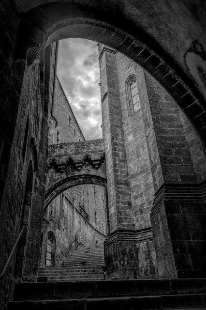 A giant multi-section stairway with tall walls towering over it leads to the top of the medieval abbey of Mont Saint Michel in France on an overcast day, in black and white. 