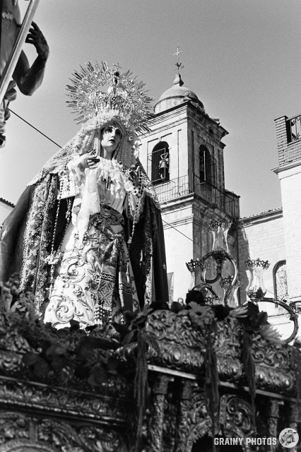A black-and-white film photo looking up at a large figure of Mary being carried on a Semana Santa float through the town streets. A church belfry is in the background.
