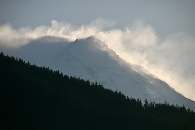 Snow-covered mountain peak with clouds of snow billowing from its slopes and a dark forest silhouette in the foreground.