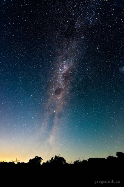 The Milky Way, with the silhouette of trees in the foreground, and the glow of an almost-full moon just below the horizon.