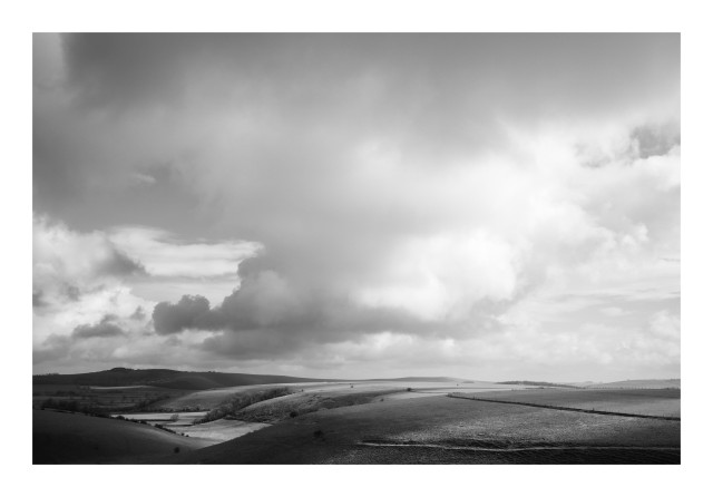 A black and white image of England's rolling hills. There are thick voluminous clouds, and dappled sunlight across the fields. 