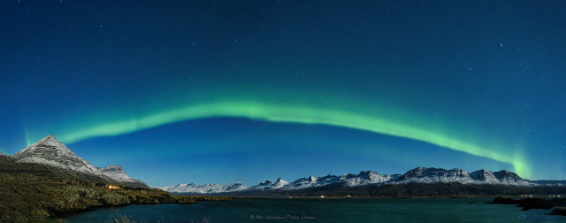 A panoramic photo of the night sky over a fjord. On the left is a lovely pyramidal mountain, and the range continues further down the waterside and across to the opposite shore on the right, all lit by moonlight which is behind the viewpoint. The sky is clear and full of stars, and a single band of bright green aurora is stretched from one side of the sky to the other. It isn't a completely smooth curve, there are flicks and kinks, and some small changes in brightness. Sharp eyes can perceive another, fainter band of green below the first. The scene is awe-inspiring and astonishingly beautiful.