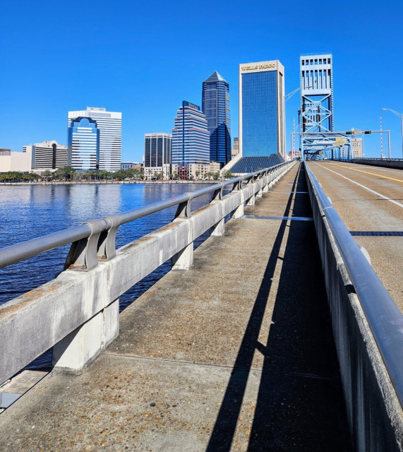 View crossing a large blue metal traffic bridge from the adjacent pedestrian crossing across a vast calm blue river with colorful skyscrapers lining the opposite shoreline beneath a brilliant clear blue sky.