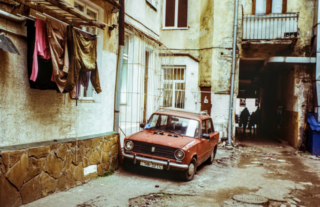 An old dark courtyard-well. A red car from the 70s is parked against the wall. The walls of the house are shabby. In some places, the plaster has fallen off and bricks are visible. On the left, laundry is hung along the wall. On the right you can see a dark passage in the house leading to the street