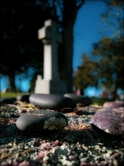 A close up of a small group of coloured stones sits atop a moss covered rock in a cemetery. In the background a blurry headstone and trees can be seen.