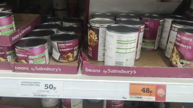 Shelf in supermarket displaying Sainsbury's own brand tins of butter beans, the small (215g) tin is 50 pence, the large (400g) is 48 pence.