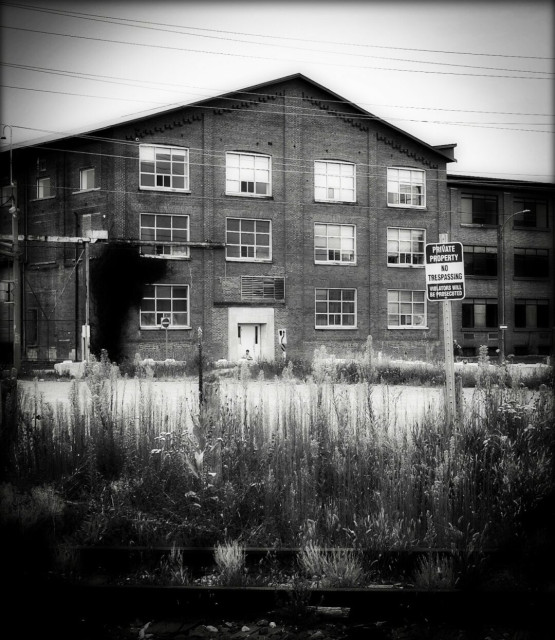 A black and white image of a large factory with many large windows. A private property no trespassing sign can be seen in the foreground along with tall grass and old railway tracks.