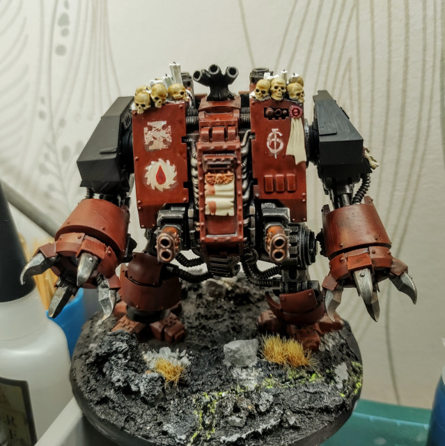 An upscaled boxy Dreadnought from Warhammer 40,000. It is painted a dark red with black and metallic details. On top of it are skulls and candles. It has claws, the better to rend heretics with.