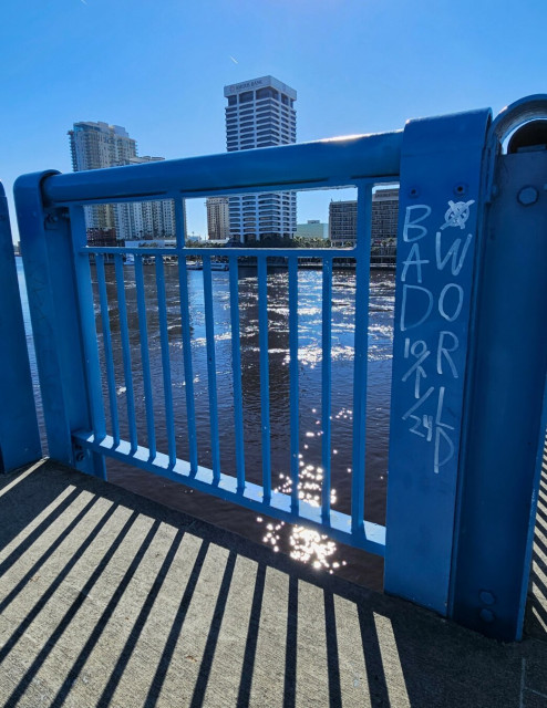 View from overlook balcony on the pedestrian crossing adjacent to the main traffic blue metal bridge. On the side in white paint is written : Bad World 10/1/24 (presumably meaning January 1, 2024 given that this image was captured in April 2024, and October hasn't occurred yet. 🤭
The railing and supports provide a cage or jail bars like shadow on the walkway. A city skyline is seen over the edge, lining the riverside with tall buildings, beneath a brilliant clear blue sky.